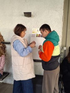 Moteris šviesia liemene padeda vaikinui su spalvinga striuke atlikti rankų darbo užduotį stovyklos terasoje.