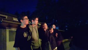 Jaunimo ir suaugusiųjų grupė šypsodamiesi žiūri į viršų vėlyvą vakarą lauke.