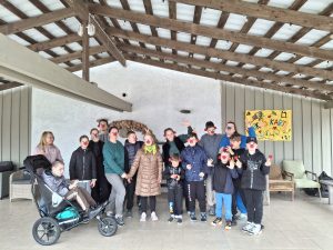 Stovyklos dalyvių ir vaikų bendra nuotrauka su raudonomis klouno nosimis terasoje prie plakato „Laikas kartu“.