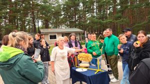 Šakočio meistrė rodo iškeptą šakotį stovyklautojams, kurie ragauja šiltus pyragaičių gabalėlius.