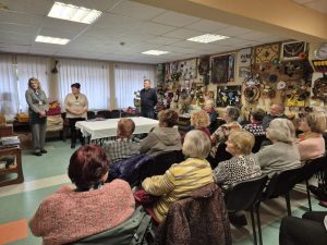 Garliavos neįgaliųjų draugijos nariai dalyvauja susitikime apie socialines paslaugas Kauno rajone. Prieš susirinkusius bendruomenės narius stovi socialinio darbo organizatorės, patalpos sienas puošia rankdarbiai.